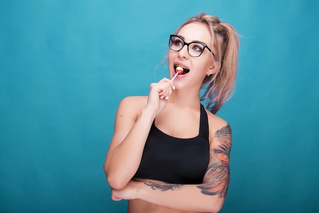 Sexy mujer rubia tatuada con una piruleta en la boca mirando su lado derecho. Foto de estudio sobre fondo azul cian