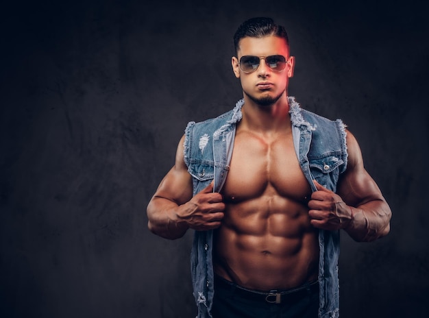 Sexy nackter modischer athletischer Mann in Jeansweste und Sonnenbrille posiert in einem Studio. Isoliert auf dunklem Hintergrund.