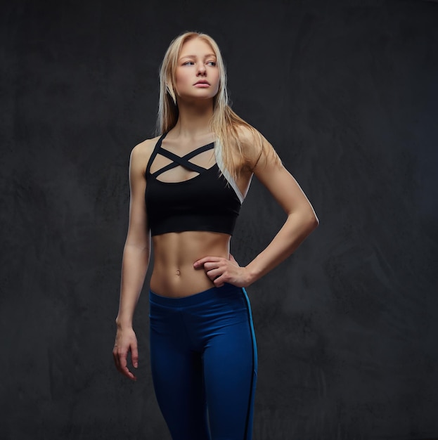 Sexy schlankes blondes Mädchen in Sportkleidung posiert in einem Studio. Getrennt auf einem dunklen Hintergrund.