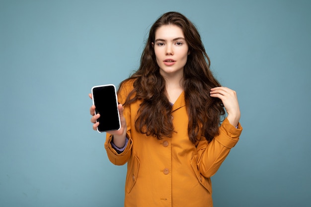 Sexy schöne junge brünette Frau mit orangefarbener Jacke isoliert auf blauem Hintergrund, die in der Hand hält und ein Mobiltelefon mit leerem Display für das Modell mit Blick auf die Kamera zeigt