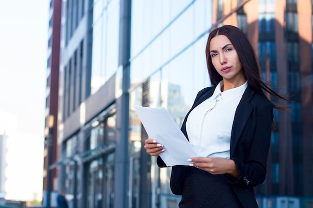 Sexy seria caliente empresaria permanente al aire libre con documentos, archivos o documentos, mirando a la cámara en ropa formal, traje, camisa blanca y chaqueta. Ocupada joven secretaria, empleado fuera de la oficina.