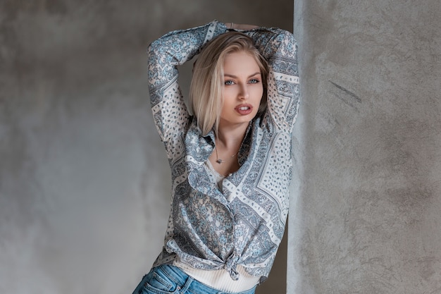 Sexy stilvolle junge blonde Frau in einem modischen Hemd mit einem Muster in einem T-Shirt in Jeans, die in einem Studio nahe einer Vintage grauen Betonwand aufwirft