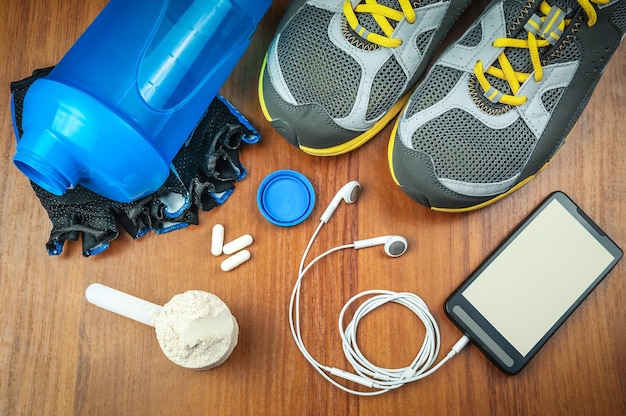 Foto shaker, proteína, calzado deportivo y smartphone con auriculares en el gimnasio.