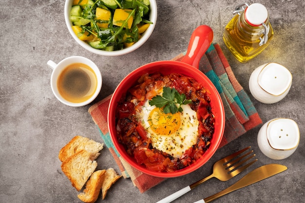 Shakshuka em uma panela vermelha, café, pão e salada em uma tigela, fundo cinza, copie o espaço