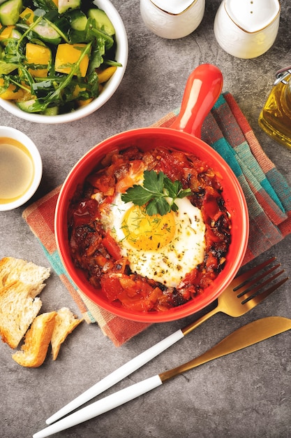 Shakshuka em uma panela vermelha sobre um fundo cinza, conceito de café da manhã com ovo frito, salada e café