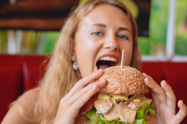 Sharming joven con cabello rubio está mordiendo una enorme hamburguesa en un café. Concepto de comida rápida. Retrato.