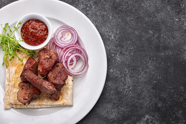 Shashlik de carne de porco, vaca, borrego, com molho, cebola e lavash, num prato branco, sobre fundo escuro