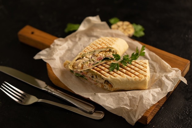 Shawarma-Brötchen mit Gemüse- und Hühnerfüllung, geschnitten auf einem Holzbrett daneben, Besteck und Knoblauch auf einem schwarzen Tisch.