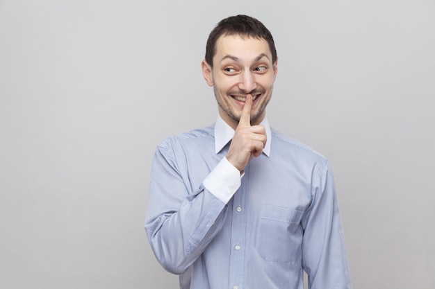 Shh, esto es secreto. Hombre de negocios de cerdas guapo divertido en camisa azul claro clásica de pie con alarma de silencio y mirando a otro lado con una gran sonrisa. tiro del estudio de interior, aislado en fondo gris.