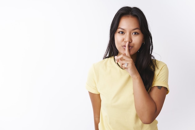 Shh halte es geheim. porträt einer mysteriösen und süßen tätowierten jungen vietnamesischen frau der 20er jahre im t-shirt, die sich vor der kamera schüttelt, um überraschung zu verbergen, zu flüstern, vor weißem hintergrund zu posieren