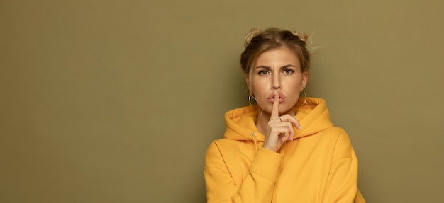 Shhh! Retrato de la muchacha bastante misteriosa en el equipo amarillo casual que muestra la muestra del silencio que sostiene el índice en fondo del color.