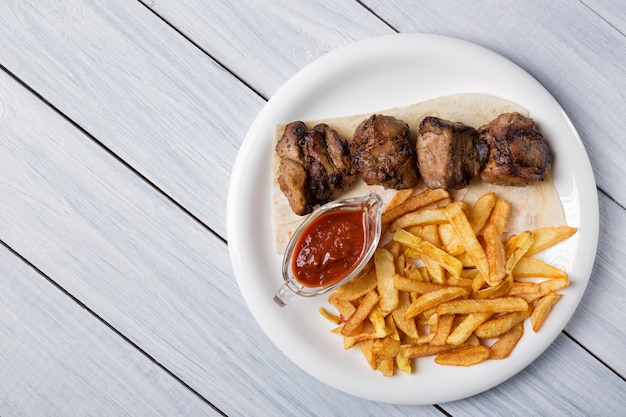 Shish kebab a la parrilla servido con papas fritas y salsa en la mesa de madera blanca