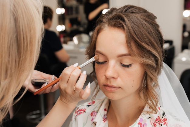 Shooting in einem Schönheitssalon. Maskenbildner trägt Make-up auf eine junge schöne Mädchenbraut auf.