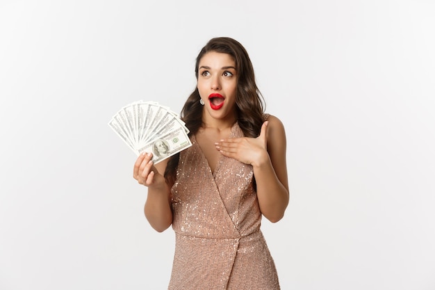 Shopping-Konzept. Bild einer überraschten und aufgeregten Frau, die Geld hält, in die obere linke Ecke starrt, erstaunt, mit Dollar im Glamour-Kleid, weißer Hintergrund stehend.