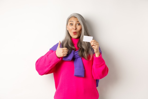 Shopping-Konzept. Lächelnde asiatische Frau mittleren Alters mit grauem Haar, die Plastikkreditkarte und Daumen hoch zeigt und Bankförderung empfiehlt, weißer Hintergrund.