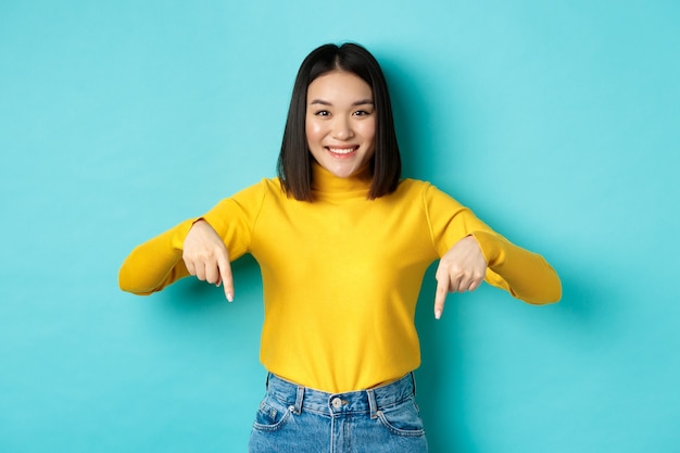 Foto shopping-konzept. schönes koreanisches mädchen mit glücklichem lächeln, das mit den fingern auf das banner zeigt und vor blauem hintergrund steht