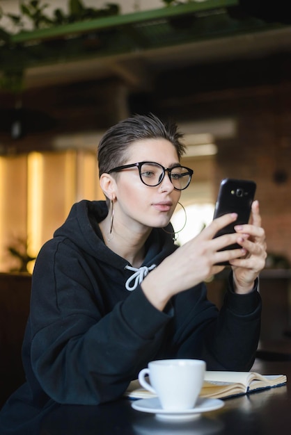 Shortcut Hipster junge Bloggerin liest Kommentare unter Post in sozialen Netzwerken auf einem modernen Telefongerät, das in der Cafeteria am Holztisch mit Buch sitzt