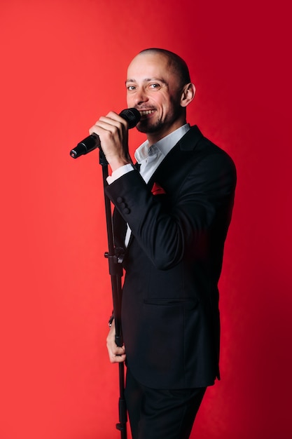 Showman-entrevistador con emociones. Un hombre joven y elegante sostiene un micrófono en una pared roja. El concepto de un showman.
