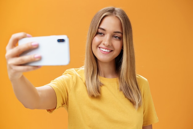 Shto cintura para arriba de la mujer saliente que sostiene el teléfono inteligente tirado en la distancia sonriendo ampliamente en la pantalla del teléfono celular mientras toma selfie