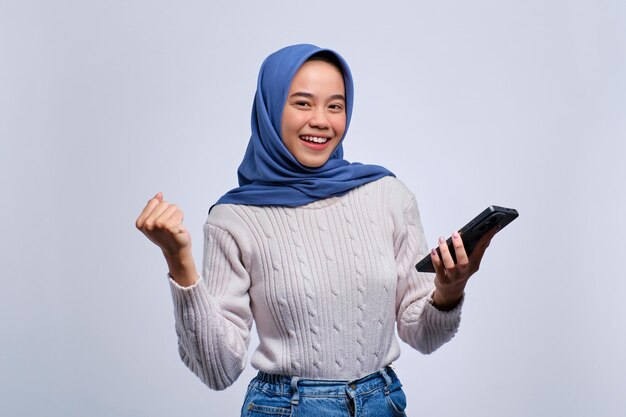 Sí, buenas noticias. Una joven asiática feliz usando un teléfono inteligente haciendo un gesto de victoria aislado sobre un fondo blanco.