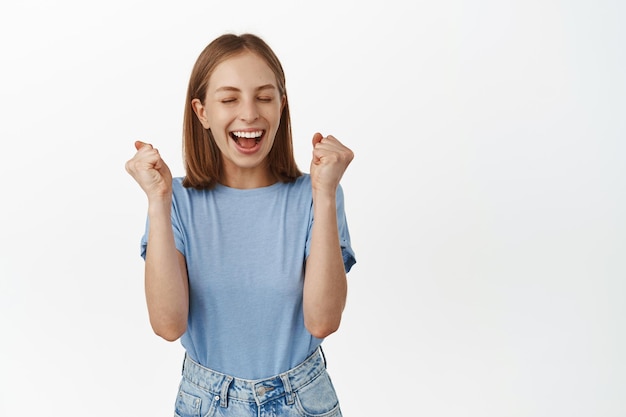 Sí, ella ganó. Una joven feliz ganando el premio y celebrando, hace puñetazos y grita de placer y alegría, logra el objetivo, triunfa, se para sobre fondo blanco.