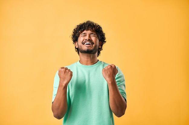 Sí Emoción de alegría celebración de la victoria Un hombre indio guapo se regocija sinceramente con los ojos cerrados y hace un gesto ganador mientras está de pie sobre un fondo amarillo