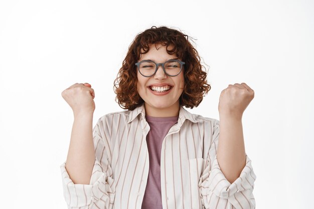 Sí, éxito. La chica moderna sonriente emocionada celebra la victoria, hace que el puño bombee para triunfar el logro de la meta, disfruta de la victoria, conviértete en ganador, de pie en blanco