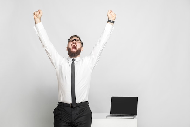 ¡Sí! Feliz, gritando, joven empresario barbudo con camisa blanca y corbata negra están de pie cerca de su lugar de trabajo y están triunfando con la boca abierta y los brazos levantados. Aislado, tiro del estudio, fondo gris