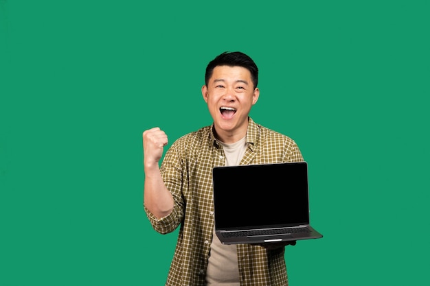 Sí, hombre coreano muy contento que sostiene una computadora portátil con pantalla en blanco y agita el puño cerrado que muestra vacío