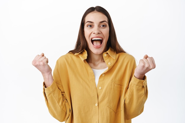 Sí, lo hice Emocionada mujer gritando ganando celebrando la victoria apretando los puños con éxito y sonriendo ampliamente gritando de alegría y felicidad triunfando de pie sobre fondo blanco