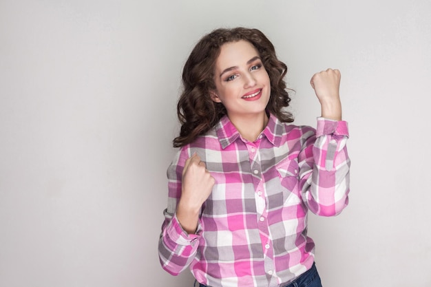 Sí lo hice. hermosa chica feliz con camisa rosa a cuadros, peinado rizado de pie, mirando a la cámara con una sonrisa dentuda y celebrando la victoria. tiro de estudio interior, aislado sobre fondo gris.