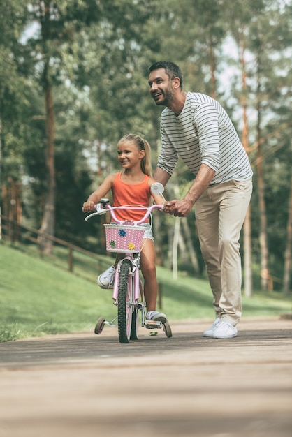 Sich in der Nähe des Vaters sicher fühlen. In voller Länge des fröhlichen Vaters, der seiner Tochter beibringt, im Park Fahrrad zu fahren?