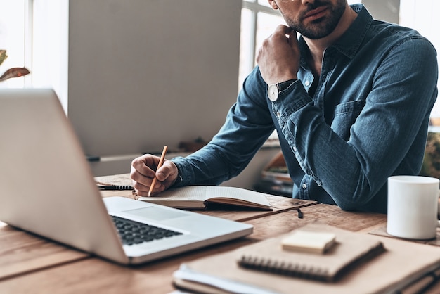 sich um das Geschäft kümmern. Nahaufnahme eines jungen Mannes, der während der Arbeit im Büro etwas aufschreibt