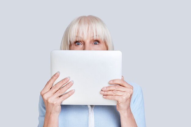 Sich verspielt fühlen. Schöne ältere Frau, die Gesicht mit digitalem Tablet bedeckt