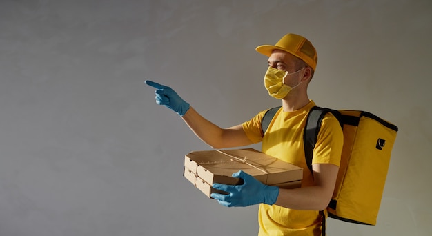 Sichere Lebensmittellieferung. Kurier in gelber Uniform, Schutzmaske und Handschuhen liefert Essen zum Mitnehmen während der Coronovirus-Quarantäne. Nach links zeigen.
