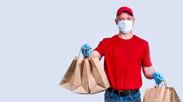 Sichere Lieferung. Kurier in roter Uniform und Schutzmaske und Handschuhen hält eine große Bestellung, viele Papiertüten, kontaktlose Zustellnahrungsmittel in Quarantäne.