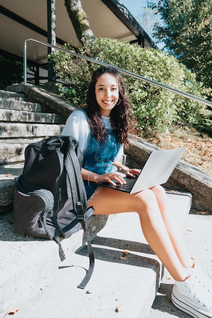 Side-shot begeisterte glückliche sorglose Studentin bereiten Projektarbeitslaptop vor, der außerhalb der Universität sitzt und modernes Outfit trägt, genießen das Klima, das zur Kamera lächelt und auf den Bildschirm schaut. Platz kopieren