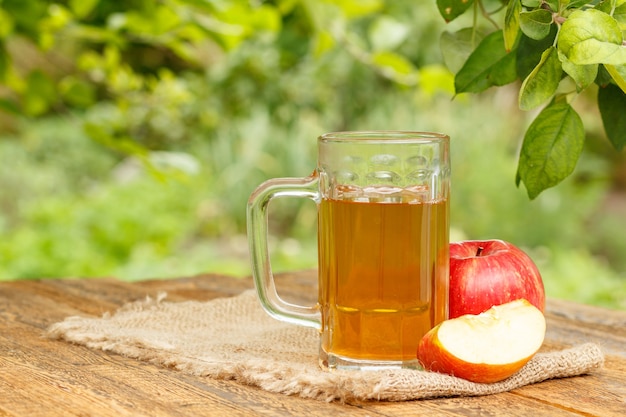 Sidra de manzana en copa de vidrio y manzanas rojas frescas sobre tablas de madera con ramas de manzano y fondo natural verde borroso a la luz del día. Profundidad de campo de Shellow. Bebidas orgánicas