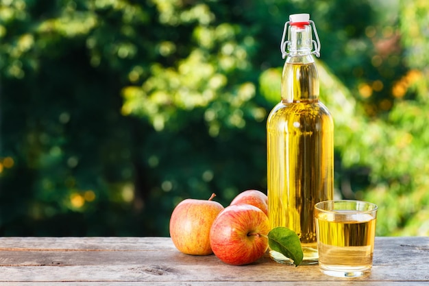 Sidra de manzana en vaso y en botella con manzanas frescas sobre una mesa de madera con fondo natural borroso