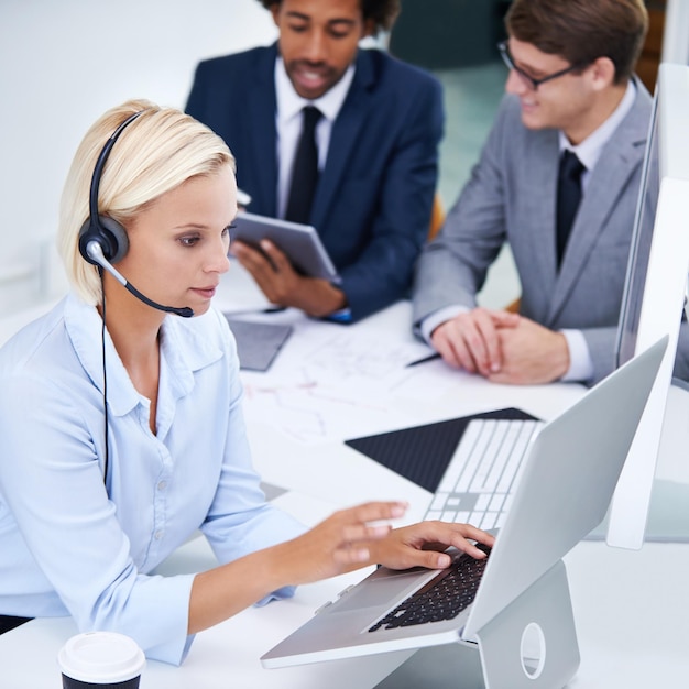 Sie hält das Büro am Laufen Aufnahme einer Geschäftsfrau, die an ihrem Laptop arbeitet, mit Kollegen im Hintergrund