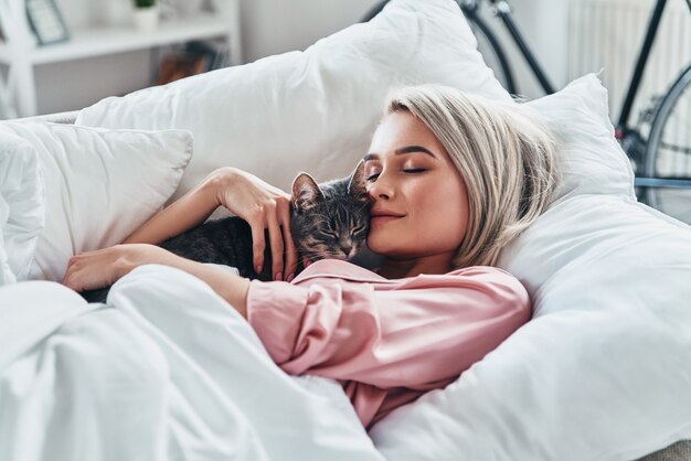 Sie liebt ihre Katze. Attraktive junge Frau, die ihre Katze hält und lächelt, während sie zu Hause im Bett liegt