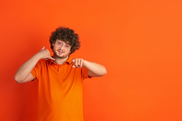 Sie müssen mich anrufen. Das monochrome Portrait des kaukasischen jungen Mannes auf oranger Wand. Schönes männliches lockiges Modell im lässigen Stil.