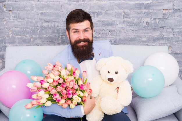 Sie verdient alles Beste Romantischer Mann mit Blumen und Teddybär sitzen auf der Couch mit Luftballons wartende Freundin Romantisches Geschenk Macho bereit romantisches Date Mann trägt Smoking Fliege halten Blumenstrauß