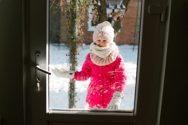 Siebenjähriges süßes Mädchen in Winterkleidung steht vor der Tür, auf der Straße mit Schnee in den Händen und schaut lächelnd ins Haus.