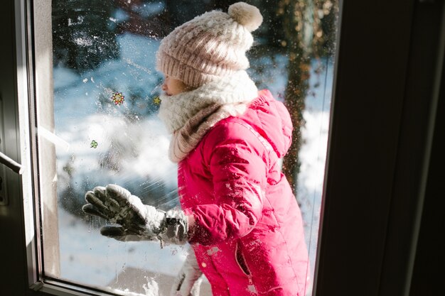 Siebenjähriges süßes Mädchen in Winterkleidung steht vor der Tür, auf der Straße mit Schnee in den Händen und schaut lächelnd ins Haus.