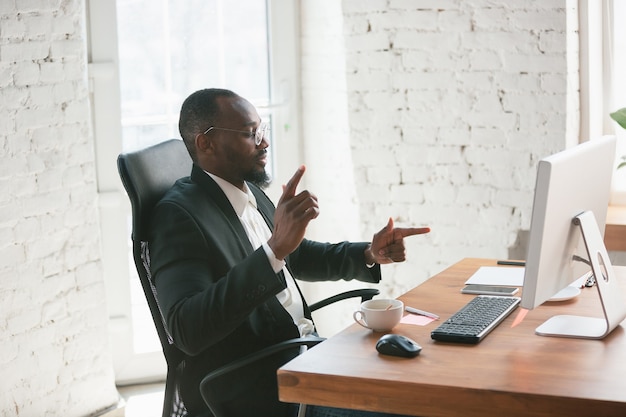 Sieg feiern. Afroamerikanischer Unternehmer, Geschäftsmann, der im Büro konzentriert arbeitet. Sieht glücklich aus, fröhlich, trägt klassischen Anzug, Jacke. Konzept der Arbeit, Finanzen, Geschäft, Erfolgsführerschaft