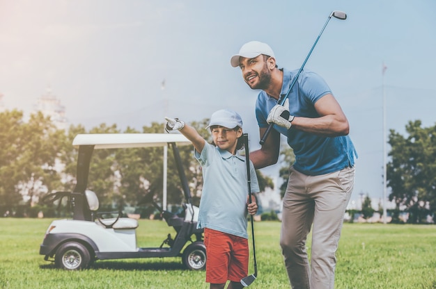 Sieh dir das an! Fröhlicher junger Mann, der seinen Sohn umarmt und wegschaut, während er auf dem Golfplatz steht