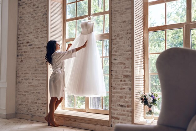 Sieht genau richtig aus. In voller Länge der schönen jungen Frau im Seidenbademantel, die ihr Hochzeitskleid berührt, während sie in der Nähe des Fensters steht