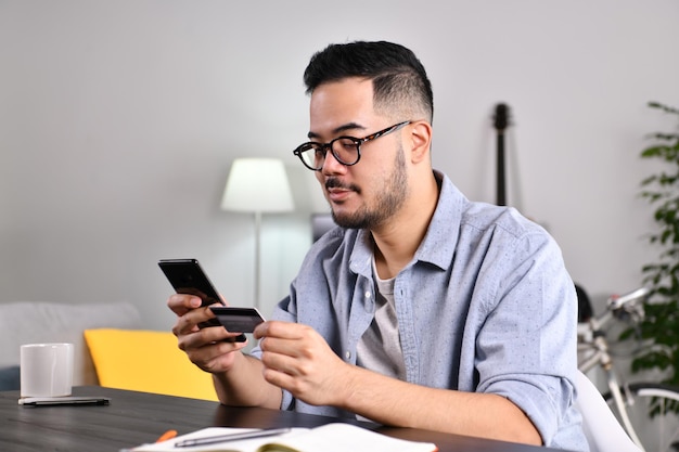 Sieht positiv aus und fühlt sich selbstbewusst. Junger asiatischer Mann mit Smartphone und Kreditkarte für Online-Shopping und Zahlung zu Hause.