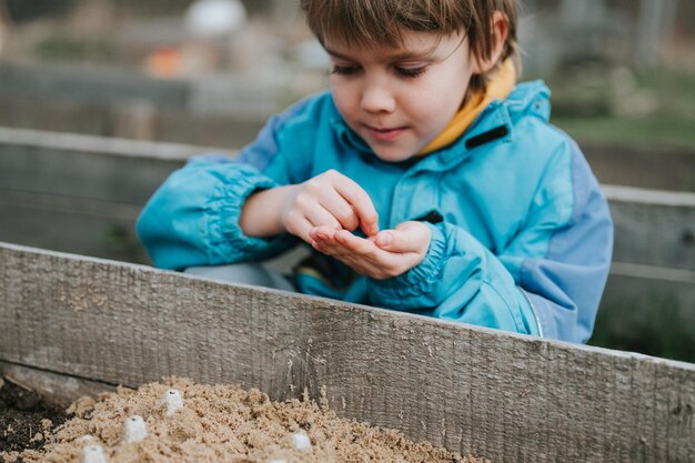 Siembra de primavera en el jardín de la granja, niño pequeño de seis años, agricultor, jardinero, plantas y siembra semillas de hortalizas en el suelo en la cama, jardinería y comienzo de la temporada de verano en el pueblo rural.